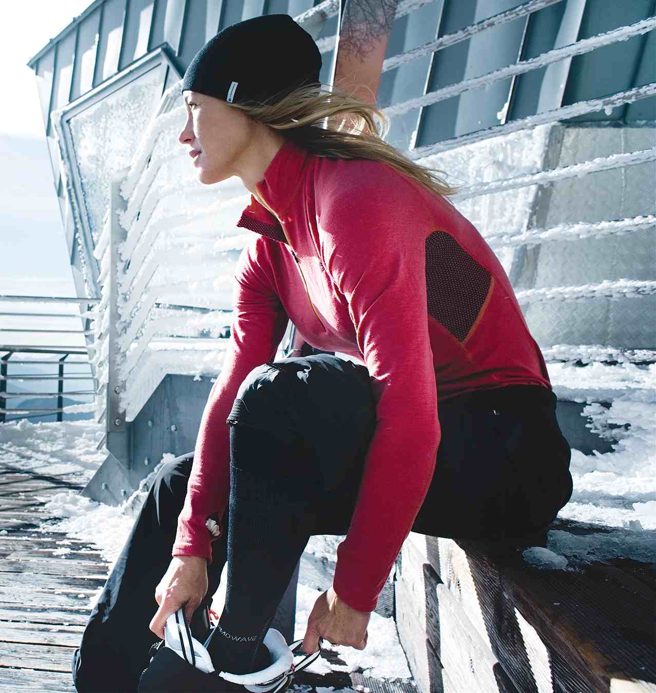 A woman wears a red thermal shirt and puts boots on her feet, surrounded by snow and mountains