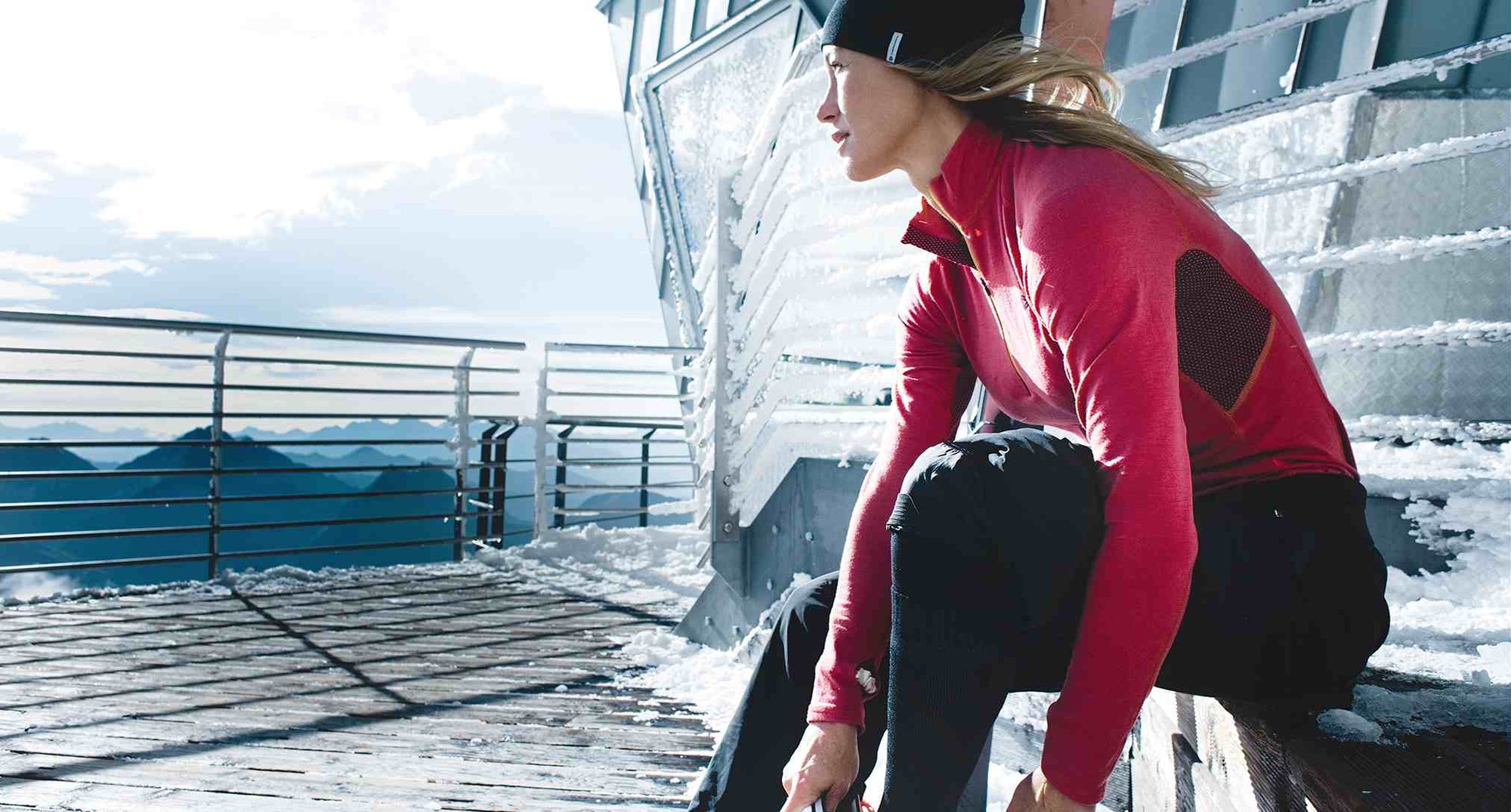 A woman wears a red thermal shirt and puts boots on her feet, surrounded by snow and mountains