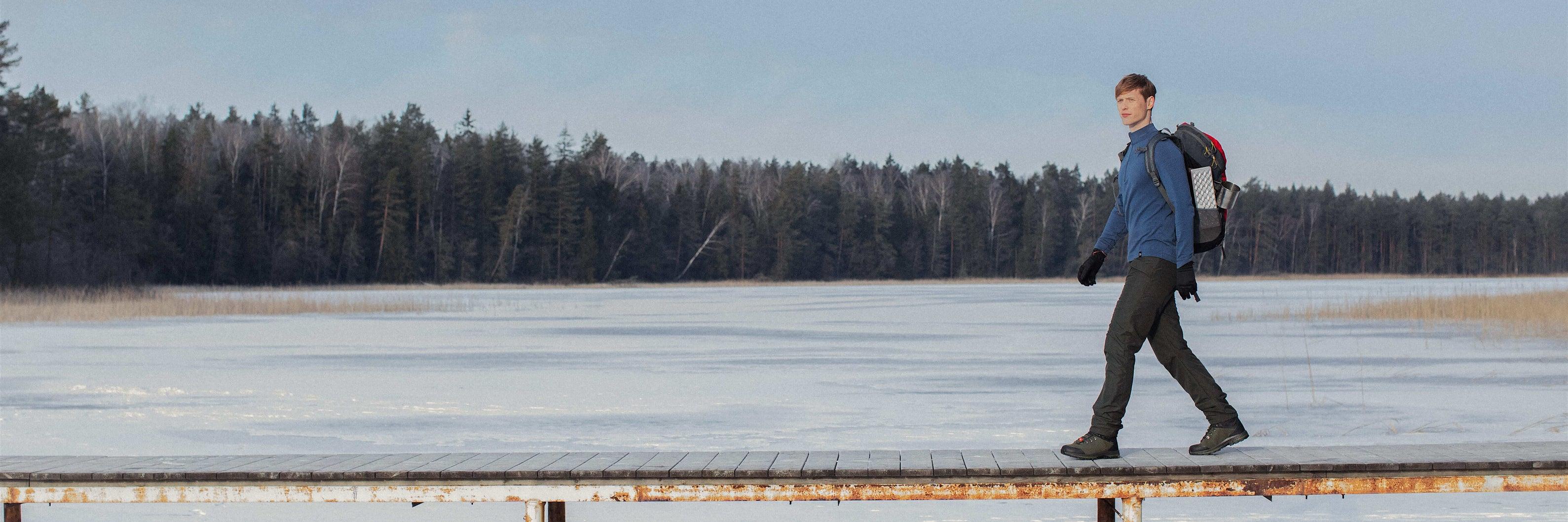 a-man-with-a-backpack-walks-on-a-bridge-over-a-frozen-lake-in-a-forest